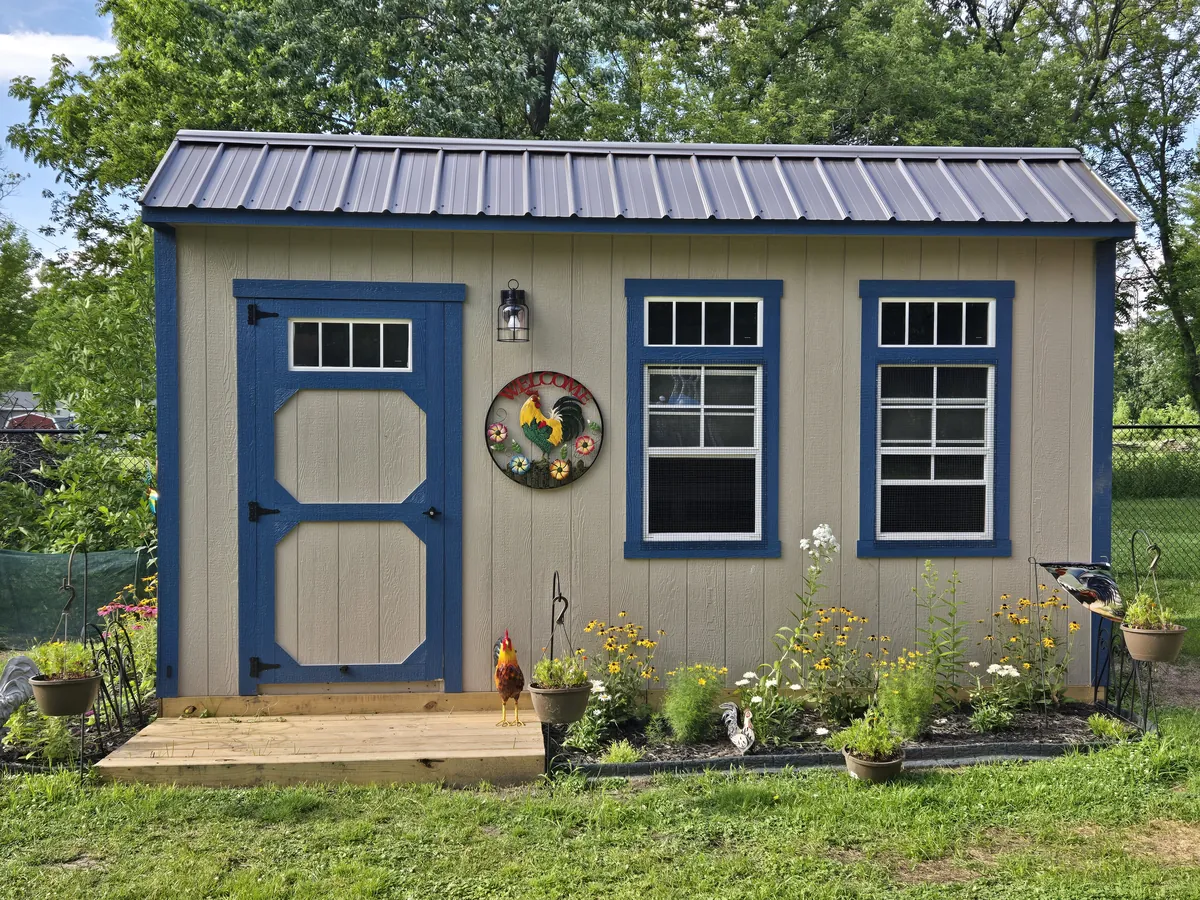 Tan garden shed with blue-trimmed door and windows, a metal roof, and flower beds in front.