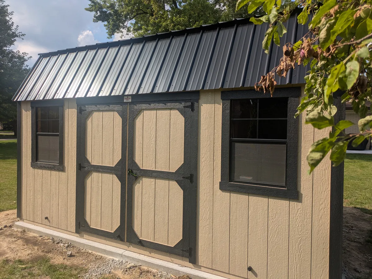 Tan lofted barn shed with dark trim, a black metal roof, and double doors.