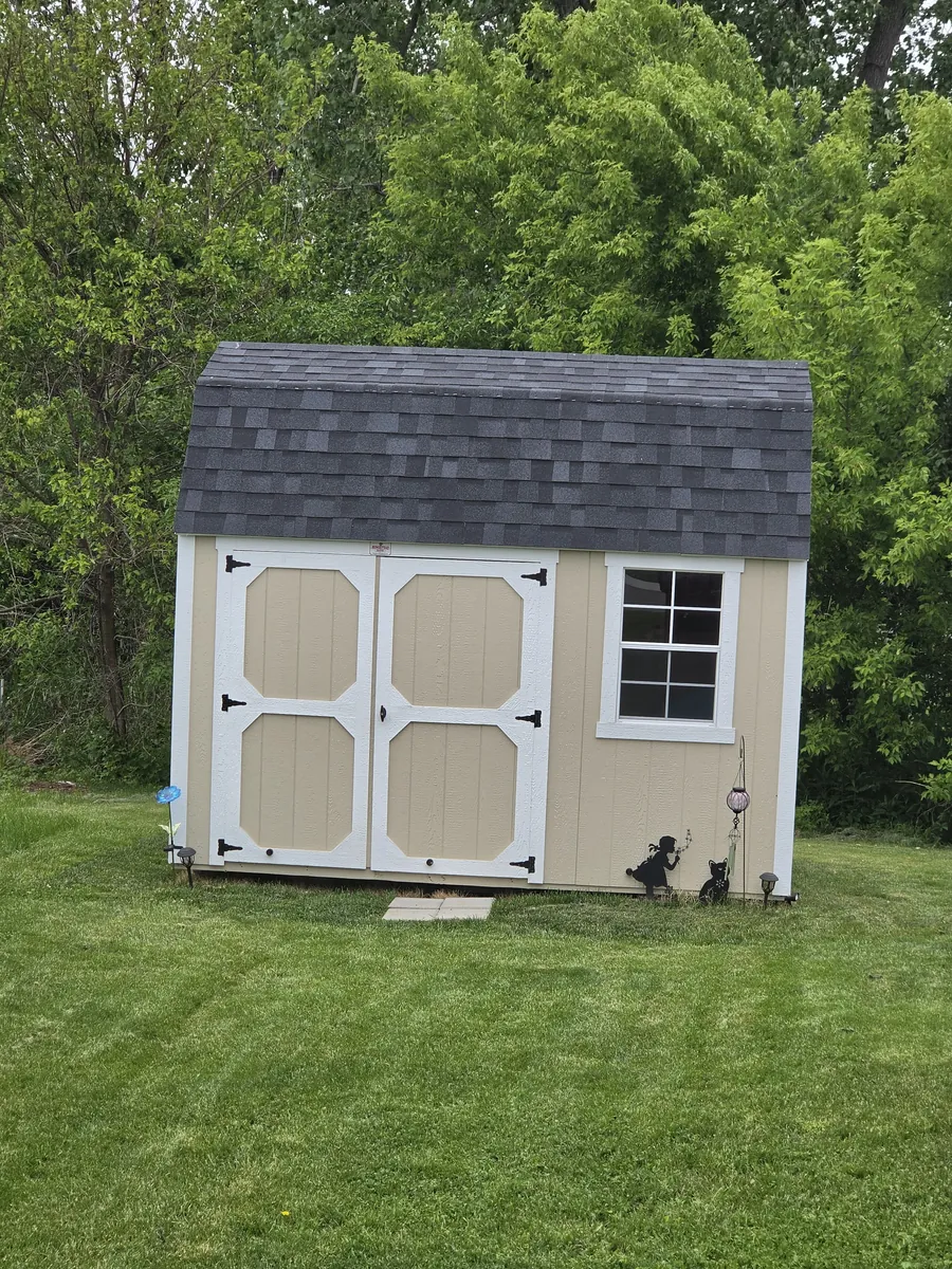 Tan lofted barn shed with white trim, double doors, and shingled gambrel roof on a lawn near trees.