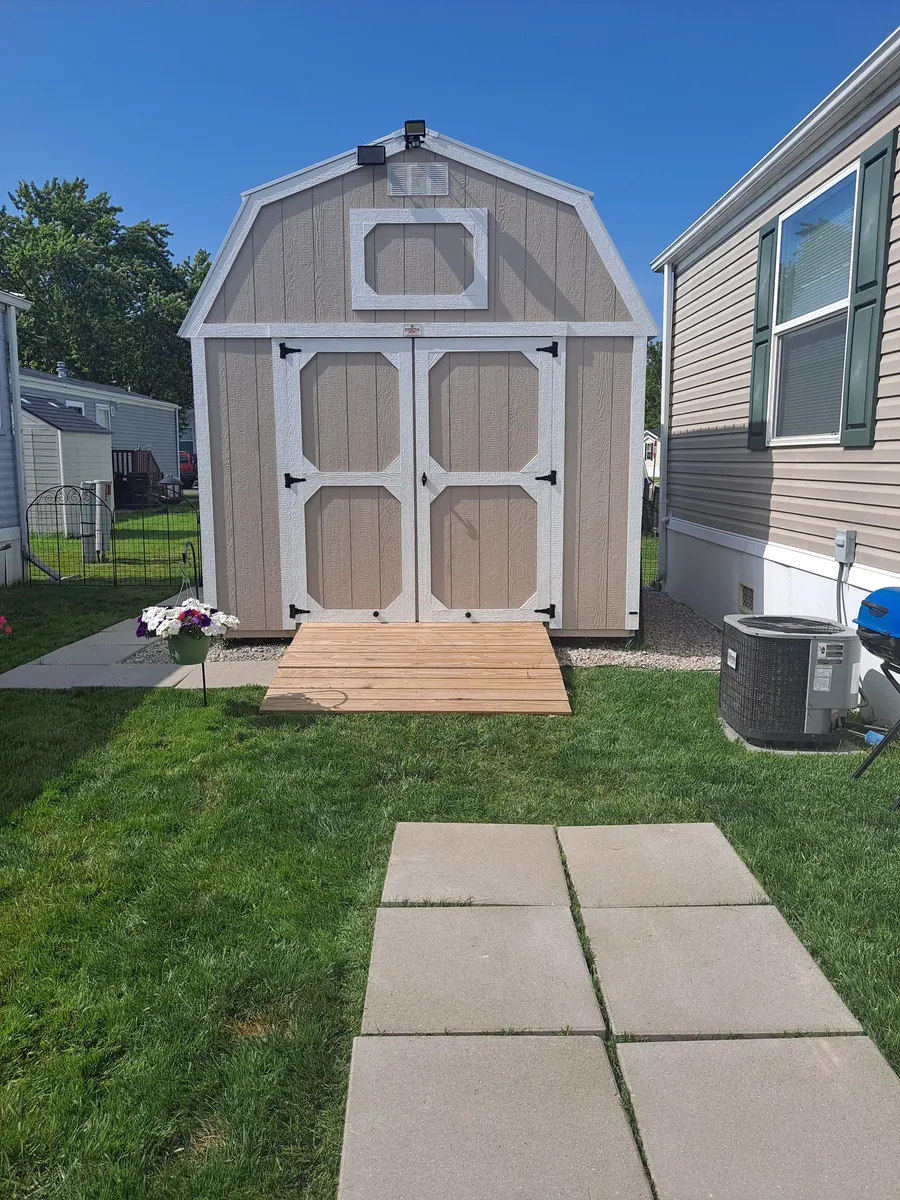 Tan lofted barn shed with white trim and a wooden ramp beside a home, with a paver walkway in front.