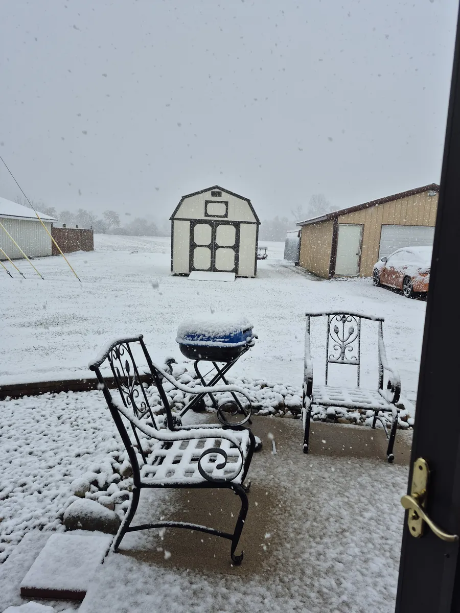 Tan lofted barn shed seen across a snow-covered yard with patio furniture during a snowstorm.