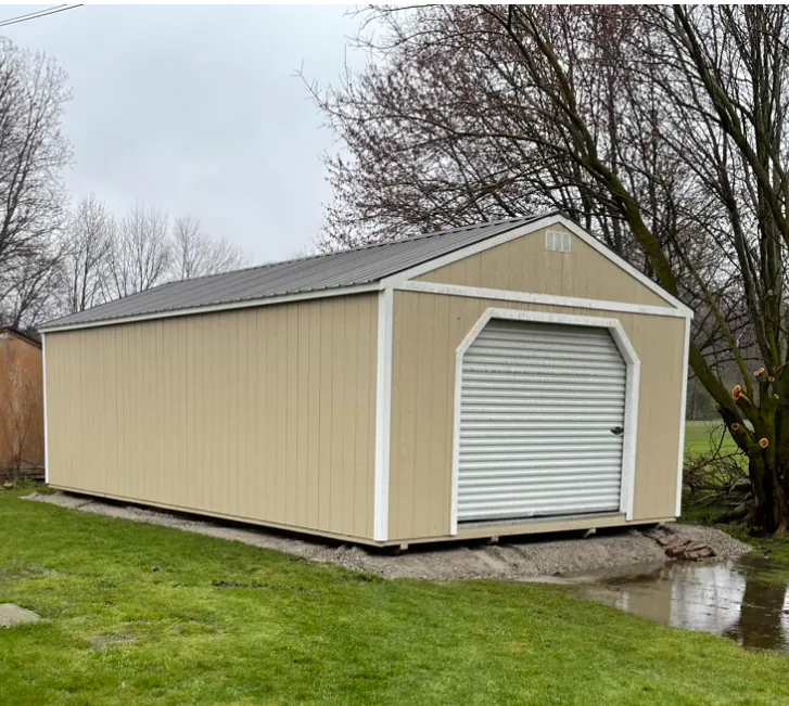 Tan portable garage with white trim, a metal roof, and a roll-up door on a gravel pad.
