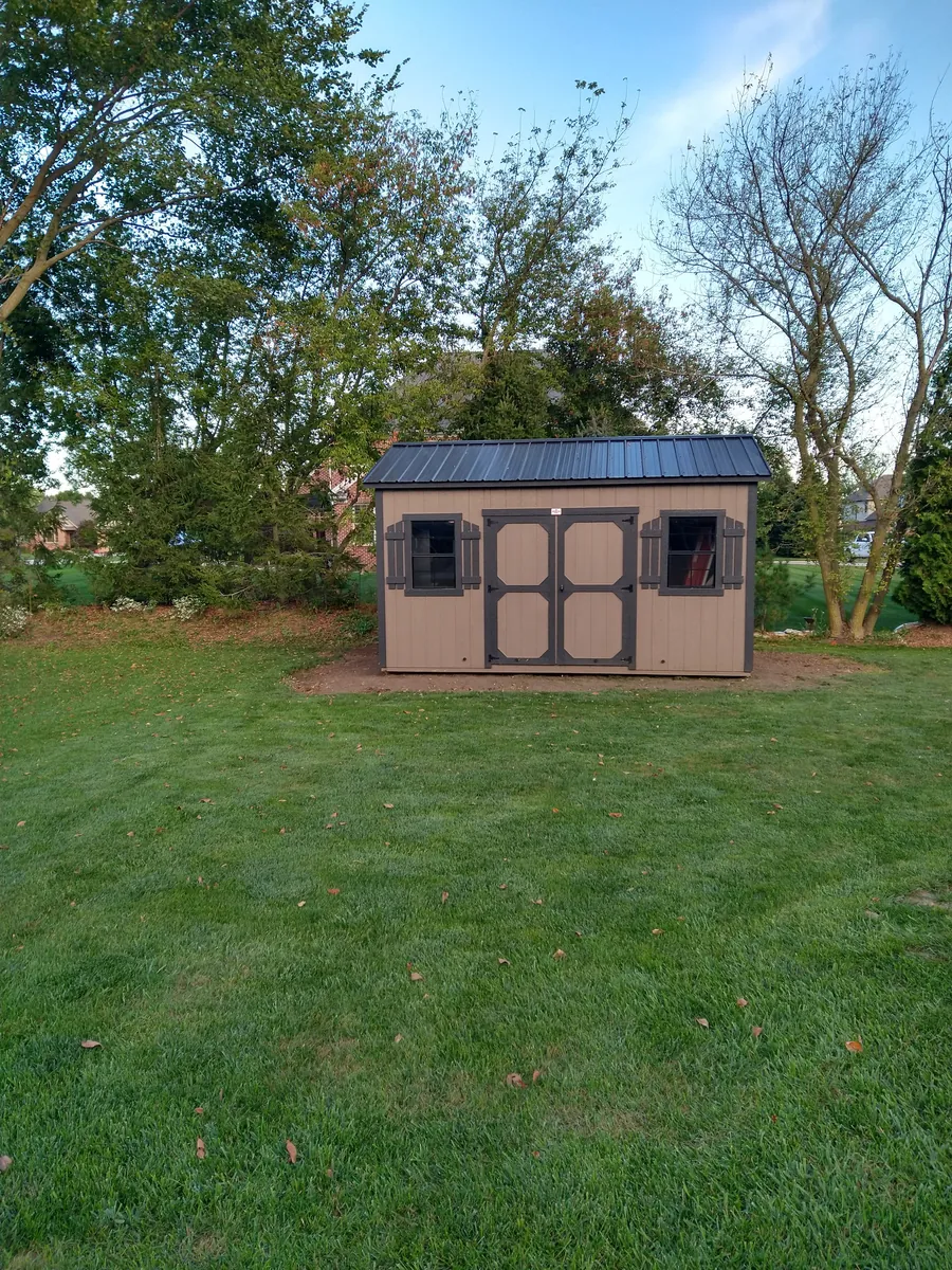 Tan shed with brown-trimmed double doors, shuttered windows, and a black metal roof on a large lawn.