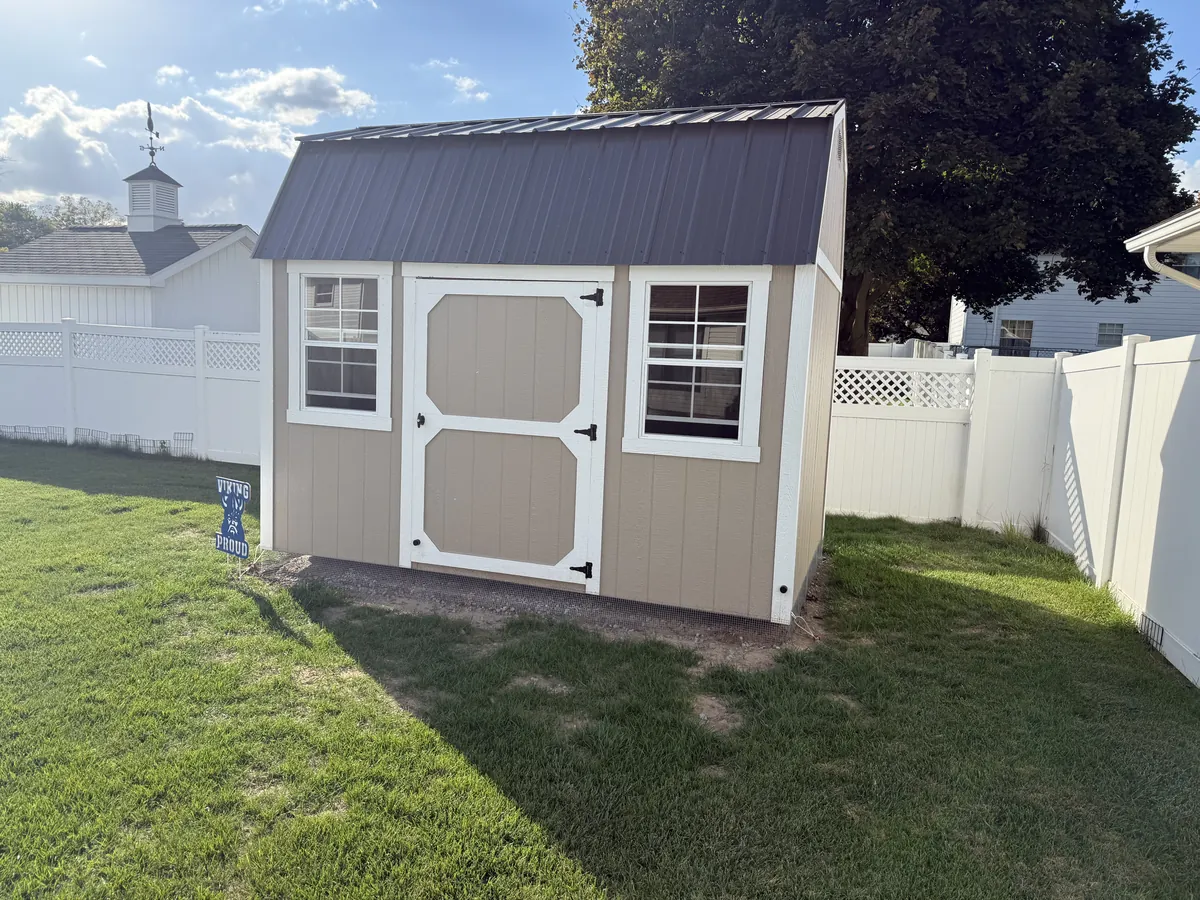 Tan shed with white trim and a dark metal roof in a backyard with white vinyl fencing.