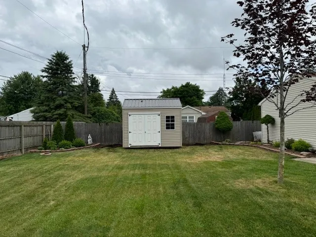 Tan utility shed with white double doors on a backyard lawn along a wooden fence line.
