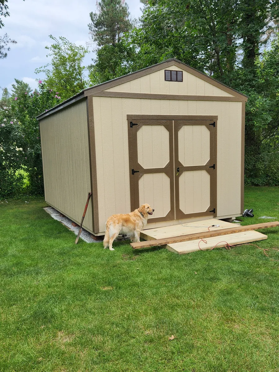 Tan utility shed with brown trim double doors on a lawn with a golden retriever nearby.