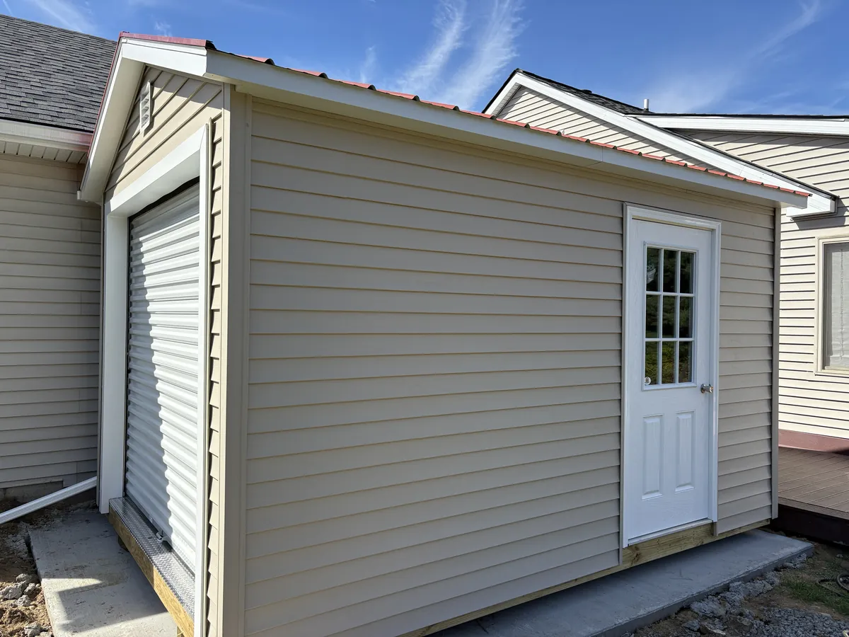 Tan vinyl-sided shed with a roll-up door and white entry door on a concrete pad between two buildings.