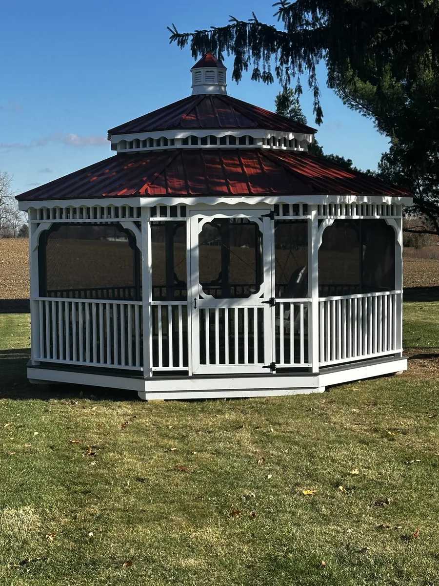 White octagonal gazebo with a red metal roof and screened sides on a lawn.