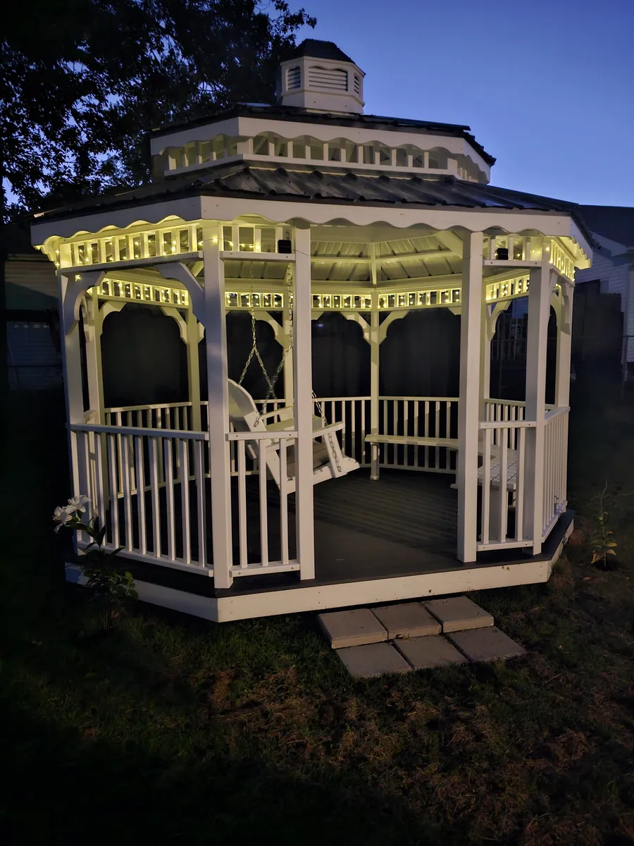 White gazebo lit with string lights at dusk, with chairs and a hanging swing inside.