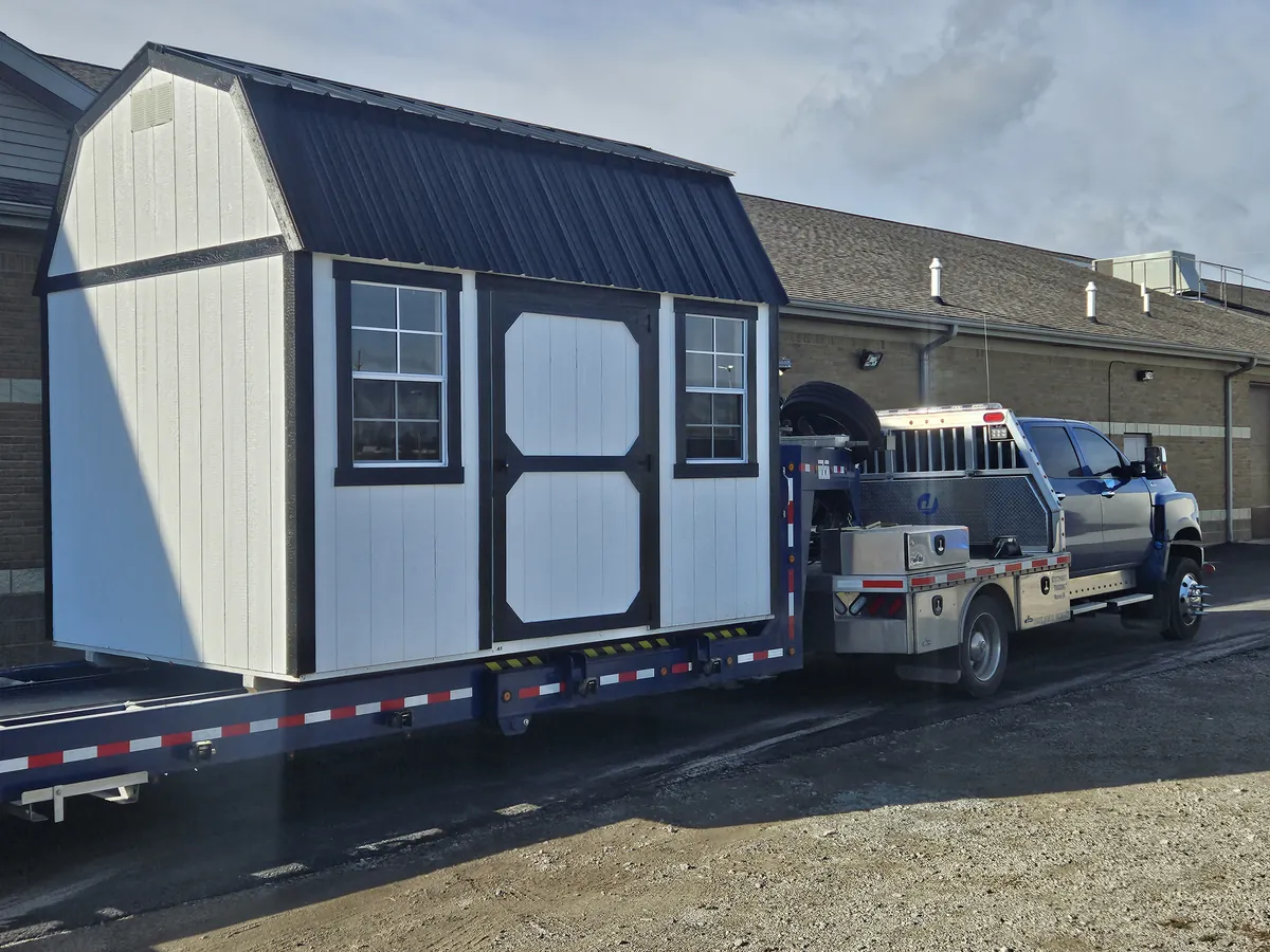 White lofted barn shed with a black roof loaded on a delivery truck and trailer.