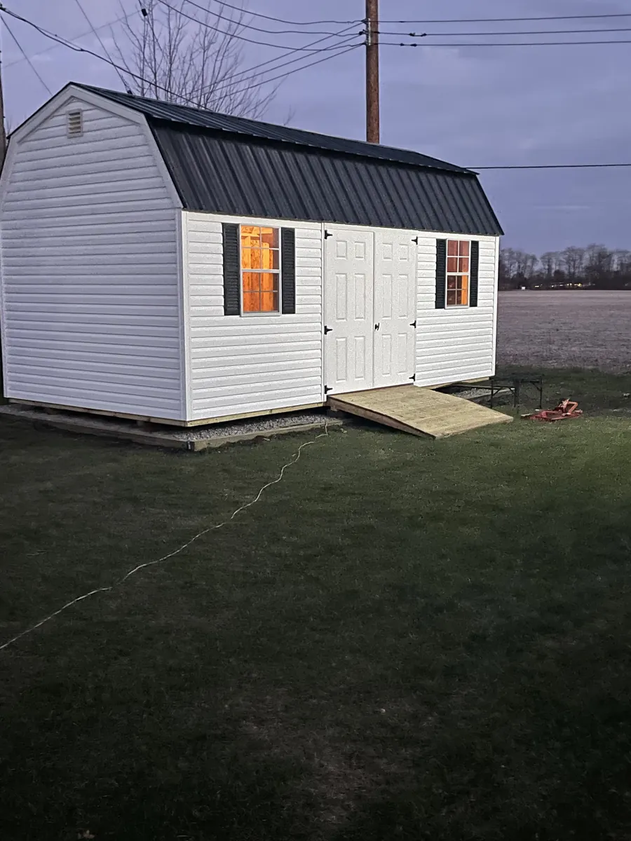 White lofted barn shed with black metal roof and lit windows at dusk beside an open field.