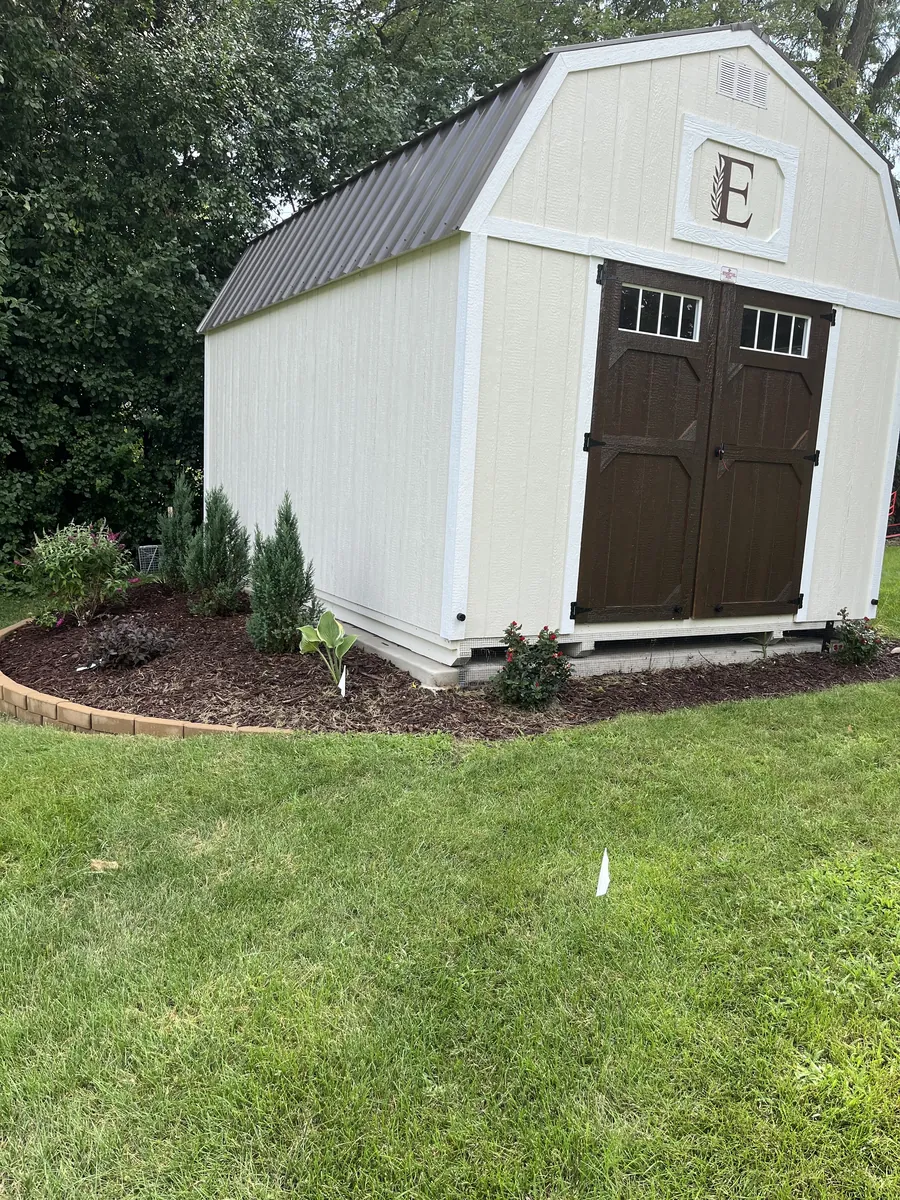 White lofted barn shed with brown double doors and a monogram, surrounded by mulched landscaping.