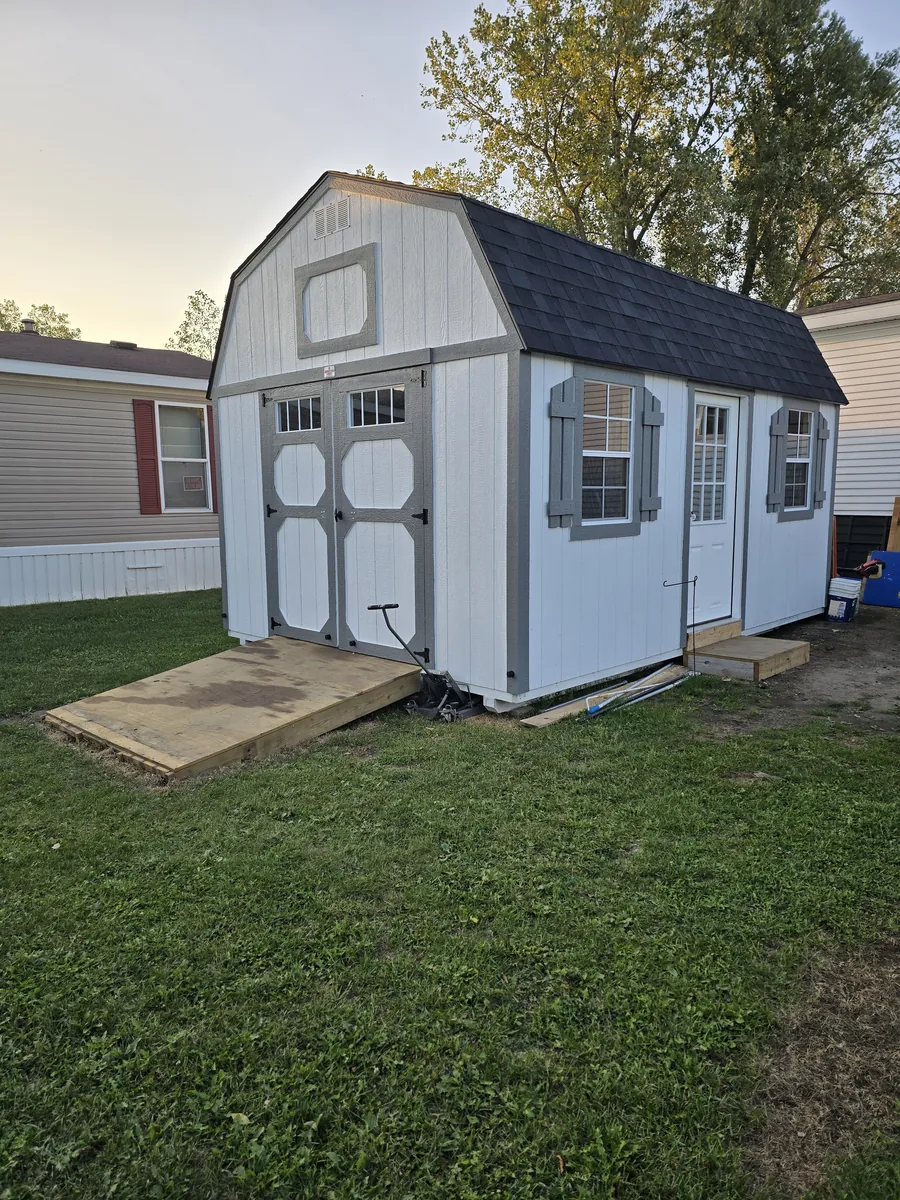 White lofted barn shed with gray trim, double doors, side entry door, and a wooden ramp in a yard.