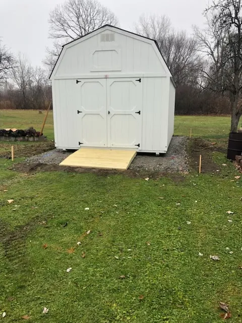 White lofted barn shed with double doors and a new wooden ramp on a gravel pad in a yard.