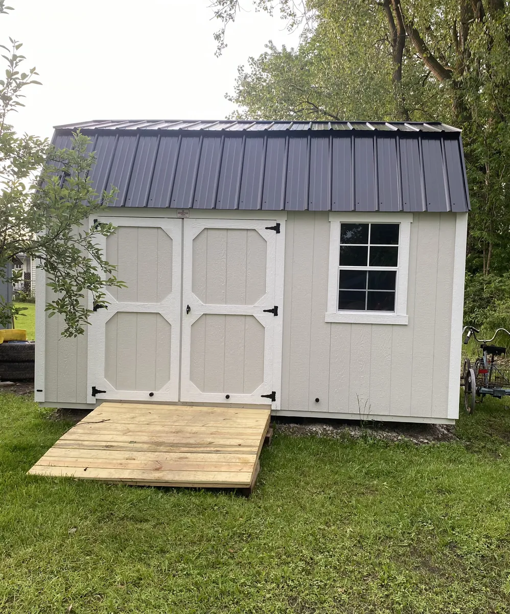 White lofted barn shed with a black metal roof, double doors, and a wooden ramp on a lawn.