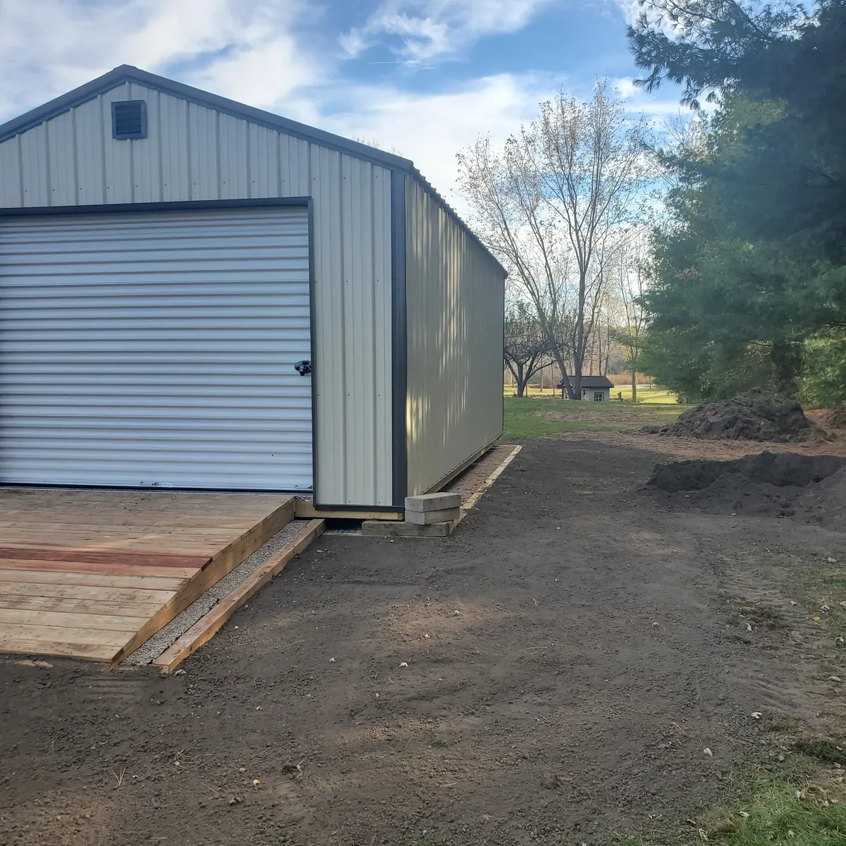 Light gray metal garage shed with a roll-up door and wooden ramp on freshly graded ground.