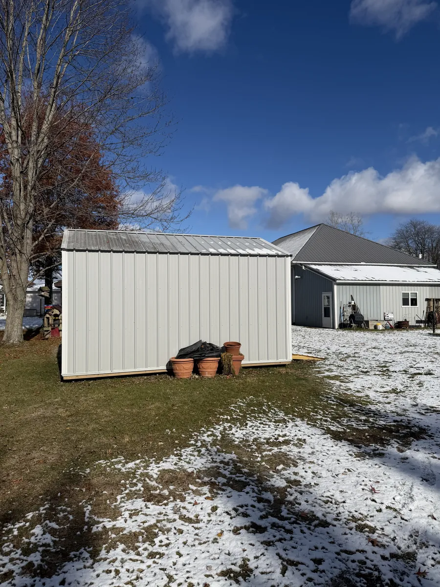 Rear view of a white shed with metal roof in a yard with light snow, with potted planters alongside.