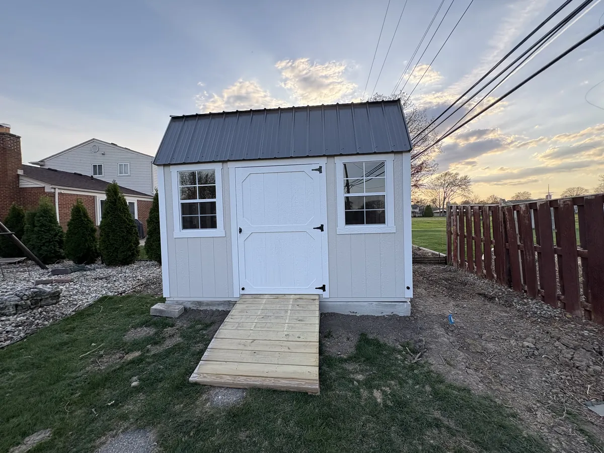 White shed with black metal roof, single door, two windows, and a wooden ramp in a backyard at sunset.