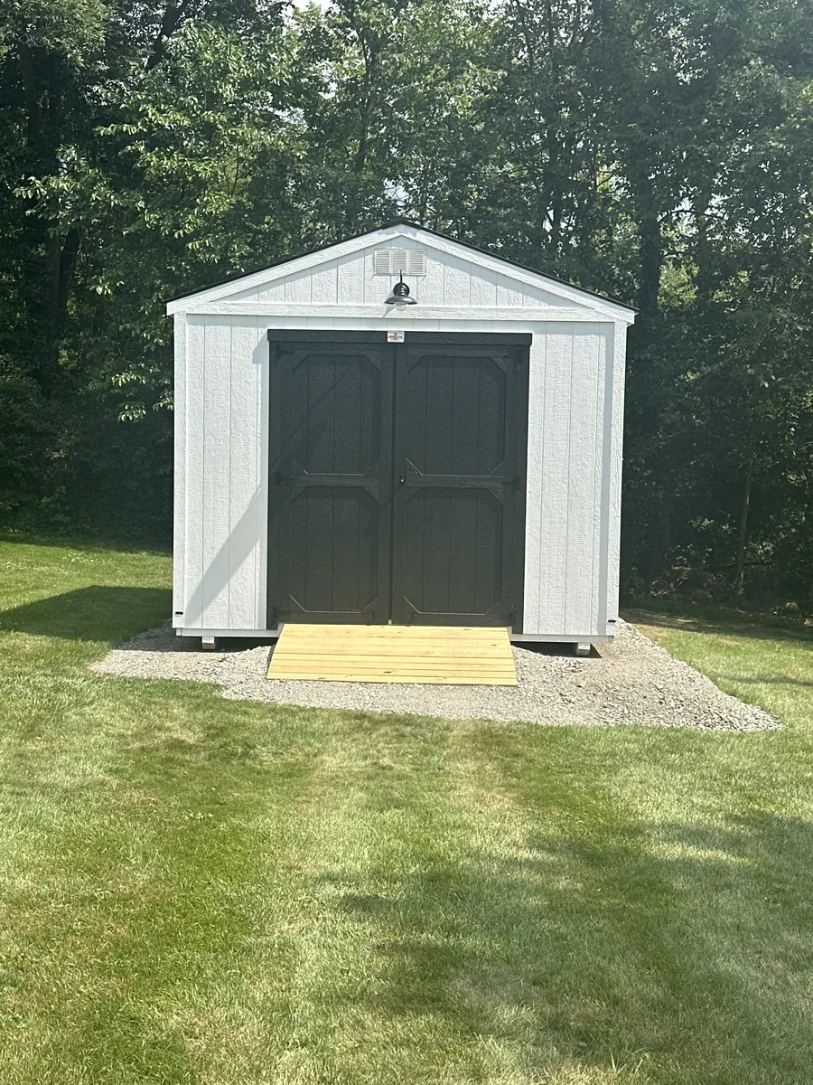 White utility shed with dark double doors and a wooden ramp on a gravel pad in a backyard.